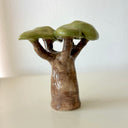 A small glazed ceramic tree figurine with a brown textured trunk and glossy green leaves, standing on a light beige surface against a neutral off-white background.