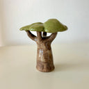 A small glazed ceramic tree figurine with a brown textured trunk and glossy green leaves, standing on a light beige surface against a white wall.