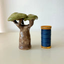 A small glazed ceramic tree figurine with a brown trunk and green leaves stands next to a spool of blue thread on a light surface against a plain white wall.