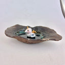 A handmade dark brown clay dish with a textured, organic shape, featuring a small white and gold ceramic mermaid animal figurine sitting in the center on a light gray background.