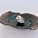 A handmade dark brown clay dish with a textured, organic shape, featuring a small white and gold ceramic mermaid animal figurine sitting in the center on a light gray background.