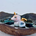A handmade dark brown clay dish with a textured, organic shape, featuring a small white and gold ceramic mermaid animal figurine sitting in the center on a light gray background.