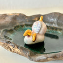 A handmade dark brown clay dish with a textured, organic shape, featuring a small white and gold ceramic mermaid animal figurine sitting in the center on a light gray background.