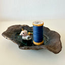 A handmade dark brown clay dish with a textured, organic shape, featuring a small white and gold ceramic mermaid animal figurine sitting in the center on a light gray background.