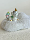 A small ceramic unicorn figurine with a pastel rainbow-colored tail and golden horn, sitting on a fluffy white cloud-shaped base against a plain light background.