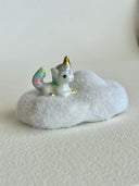 A small ceramic unicorn figurine with a pastel rainbow-colored tail and golden horn, sitting on a fluffy white cloud-shaped base against a plain light background.