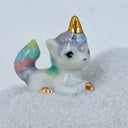 A small ceramic unicorn figurine with a pastel rainbow-colored tail and golden horn, sitting on a fluffy white cloud-shaped base against a plain light background.