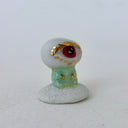 A small decorative figurine of a character with one large eye, a red pupil and a green cylindrical body. It is positioned on a white cloud-like base.