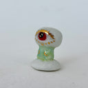 A small decorative figurine of a character with one large eye, a red pupil and a green cylindrical body. It is positioned on a white cloud-like base.