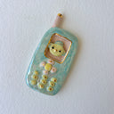 A handmade ceramic mobile phone charm or pendant in light blue and white marbled glaze, featuring a yellow cartoon face in the screen window and gold-plated button details, shown on a textured white background.