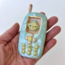 A handmade ceramic mobile phone pendant in light blue and white marbled glaze, featuring a yellow cartoon face in the screen window and gold-plated button details, shown on a textured white background.