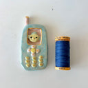 A ceramic phone-shaped object with a small bird figure inside the screen, shown next to a spool of blue thread on a plain white surface.
