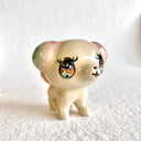 Ceramic sculpture of a small tan dog with a small body and large head, with glittery anime style eyes and green and pink ears.