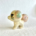 Ceramic sculpture of a small tan dog with a small body and large head, with glittery anime style eyes and green and pink ears. It has a curled tail like a squirrel.