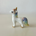 A small glossy white unicorn figurine with gold accents and pastel blue, purple, and yellow iridescent detailing on its mane, tail, and hooves, standing on a light beige surface against a neutral light gray background.