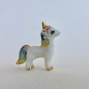 A small glossy white unicorn figurine with gold accents and pastel blue, purple, and yellow iridescent detailing on its mane, tail, and hooves, standing on a light beige surface against a neutral light gray background.