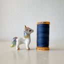 A small glossy white unicorn figurine with gold accents and pastel blue, purple, and yellow iridescent detailing on its mane, tail, and hooves, standing on a light beige surface against a neutral light gray background.