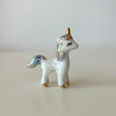 A small glossy white unicorn figurine with gold accents and pastel blue, purple, and yellow iridescent detailing on its mane, tail, and hooves, standing on a light beige surface against a neutral light gray background.