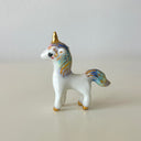 A small glossy white unicorn figurine with gold accents and pastel blue, purple, and yellow iridescent detailing on its mane, tail, and hooves, standing on a light beige surface against a neutral light gray background.