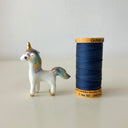 A small glossy white unicorn figurine with gold accents and pastel blue, purple, and yellow iridescent detailing on its mane, tail, and hooves, standing on a light beige surface against a neutral light gray background.
