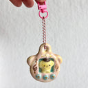 Ceramic sculpture of a small Tamagotchi doll, with 3 buttons and Pompompurin in the middle as the main character. It hangs from a pink chain.