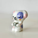 Small glossy ceramic sculpture of a white dog with pink and purple spots. It sits on its knees like a person and holds a cup of matcha.