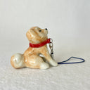 Glossy ceramic sculpture of a brown and white shiba inu sitting on the ground and wearing a red collar with a leash attached. It sits in apparent resistance.