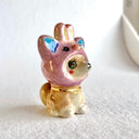 Glossy ceramic sculpture of a brown Shiba Inu sitting on the ground with glittery anime eyes. It wears a pink bunny hood hat.