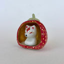 A small white ceramic fox figurine seated inside a strawberry-shaped holder with a red exterior, gold polka dots, and a pale green stem, photographed on a plain light background.
