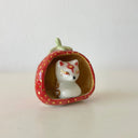A small white ceramic fox figurine seated inside a strawberry-shaped holder with a red exterior, gold polka dots, and a pale green stem, photographed on a plain light background.