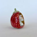 A small white ceramic fox figurine seated inside a strawberry-shaped holder with a red exterior, gold polka dots, and a pale green stem, photographed on a plain light background.