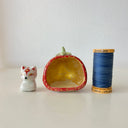A small white ceramic fox figurine seated inside a strawberry-shaped holder with a red exterior, gold polka dots, and a pale green stem, photographed on a plain light background.