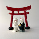 Two small decorative figurines in front of a red torii gate: one white fox-like figure with red markings and one fox-like figure  in a black robe with a gold chest emblem, set on a light gray surface against a white background.