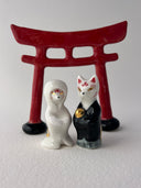 Two small decorative figurines in front of a red torii gate: one white fox-like figure with red markings and one fox-like figure  in a black robe with a gold chest emblem, set on a light gray surface against a white background.