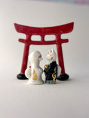 Two small decorative figurines in front of a red torii gate: one white fox-like figure with red markings and one fox-like figure  in a black robe with a gold chest emblem, set on a light gray surface against a white background.
