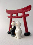 Two small decorative figurines in front of a red torii gate: one white fox-like figure with red markings and one fox-like figure  in a black robe with a gold chest emblem, set on a light gray surface against a white background.