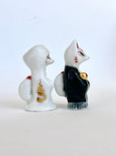 Two small decorative figurines in front of a red torii gate: one white fox-like figure with red markings and one fox-like figure  in a black robe with a gold chest emblem, set on a light gray surface against a white background.