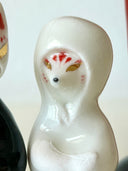 Two small decorative figurines in front of a red torii gate: one white fox-like figure with red markings and one fox-like figure  in a black robe with a gold chest emblem, set on a light gray surface against a white background.