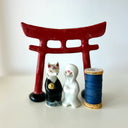 Two small decorative figurines in front of a red torii gate: one white fox-like figure with red markings and one fox-like figure  in a black robe with a gold chest emblem, set on a light gray surface against a white background.