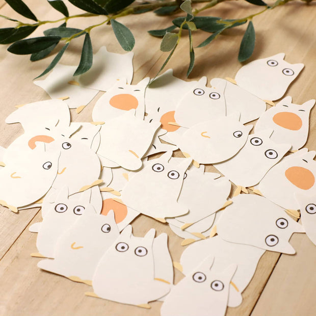 White Totoro-shaped paper cutouts with orange accents on a wooden surface with green leaves.