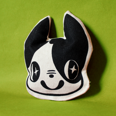Small pillow, shaped like the head of a Boston Terrier dog. It has cartoonish sparkling eyes and a happy smile and looks off to the side.