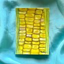 Artwork depicting a close-up yellow corn cob, placed on a shiny turquoise fabric background. The artwork has visible brushstrokes and a small artist signature in the lower right corner.