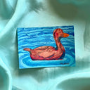 A small painting of a reddish-brown duck floating on blue water, placed on draped light aqua fabric.
