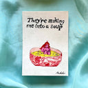 Artwork with the text 'They're making me into a soup' and a watercolor illustration of a bowl with a small purple figure, placed on a light blue satin fabric background.