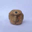 A hand-carved wood sculpture of a stylized face with a prominent bulbous nose and dark painted details, with a large 3rd eye atop its head, set against a plain light blue background.