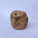 A hand-carved wood sculpture of a stylized face with a prominent bulbous nose and dark painted details, with a large 3rd eye atop its head, set against a plain light blue background.