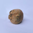 A hand-carved wood sculpture of a stylized face with a prominent bulbous nose and dark painted details, with a large 3rd eye atop its head, set against a plain light blue background.