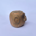 A hand-carved wood sculpture of a stylized face with a prominent bulbous nose and dark painted details, with a large 3rd eye atop its head, set against a plain light blue background.