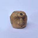 A hand-carved wood sculpture of a stylized face with a prominent bulbous nose and dark painted details, with a large 3rd eye atop its head, set against a plain light blue background.