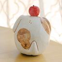 Clay sculpture of a round character, modeled to look like a dog without any arms or legs. Atop its head is an apple.